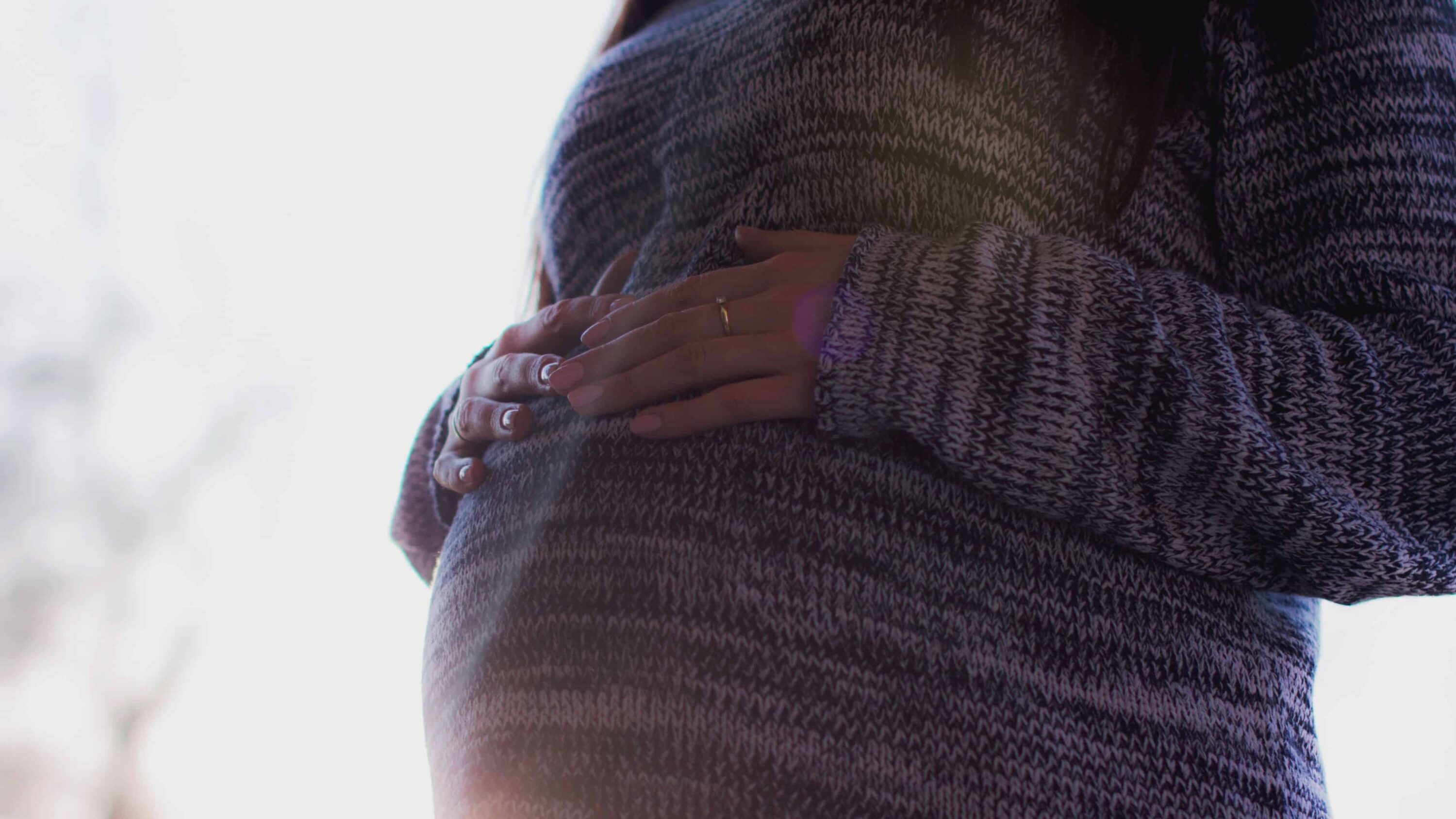 Pregnant woman in purple sweater and wedding ring resting her hands on her belly